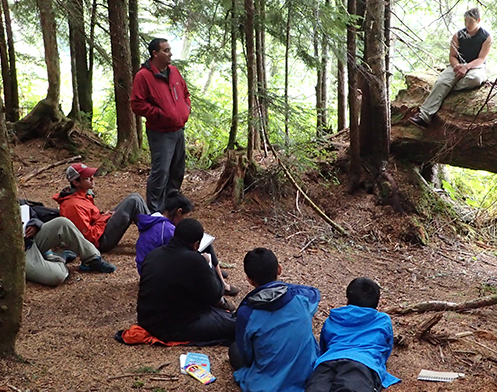 A group of young people sit in a wooded area as an adult shares his knowledge of nature